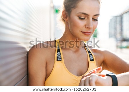 Image of caucasian woman in sportswear looking at smartwatch while walking outdoors in sunny morning
