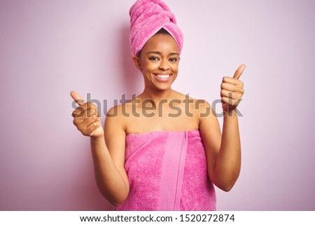 African american woman wearing shower towel after bath over pink isolated background success sign doing positive gesture with hand, thumbs up smiling and happy. Cheerful expression and winner gesture.