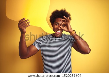 Young african american man holding speech bubble standing over isolated yellow background with happy face smiling doing ok sign with hand on eye looking through fingers