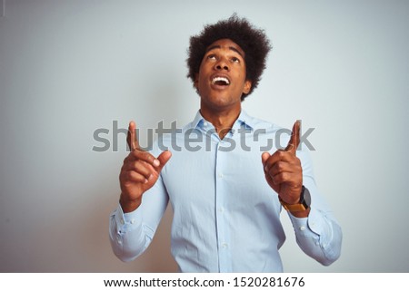 Young african american man with afro hair wearing elegant shirt over isolated white background amazed and surprised looking up and pointing with fingers and raised arms.