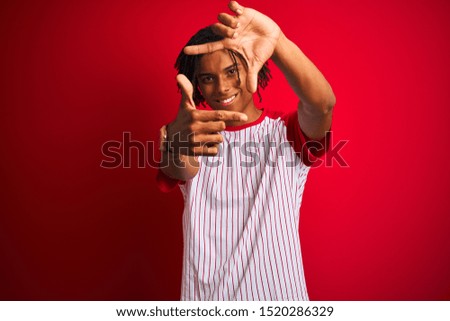 Afro man with dreadlocks wearing striped t-shirt standing over isolated red background smiling making frame with hands and fingers with happy face. Creativity and photography concept.