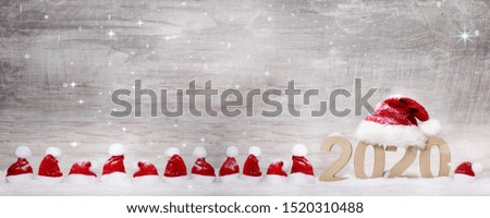 the numbers for the year 2020 stand in the deep snow with a big red santa claus hat on it, and many little santa hats in a row next to that on a wooden textured background with stars