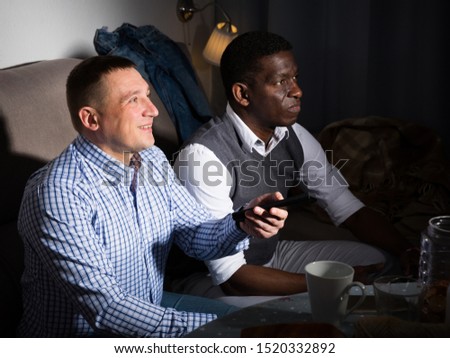 Two men resting together at home, watching tv while sitting on sofa 