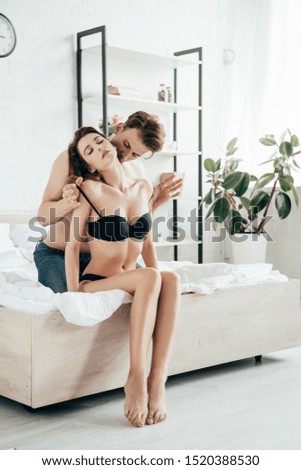 handsome boyfriend hugging and kissing attractive girlfriend in black underwear in bed