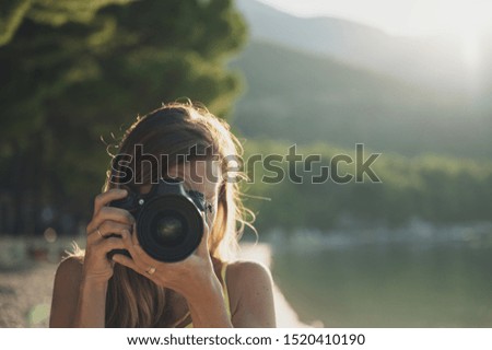 Young woman taking a photo directly at the camera with sea and beach in background.