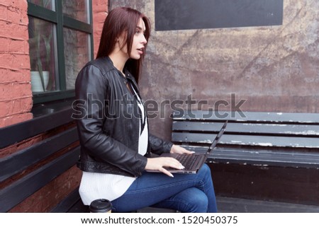 Red-haired girl in a black leather jacket works at a laptop sitting on a bench in the street