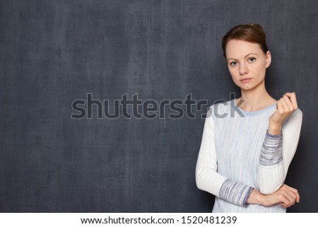 Studio half-length portrait of focused thoughtful caucasian girl wearing jumper, looking attentively and seriously at camera, standing in waiting position over gray background, copy space on left