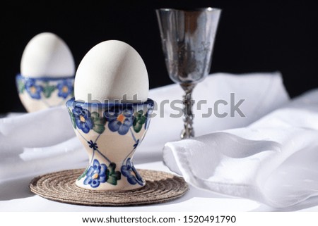Still life of two white eggs in egg stands with white napkins near it and sterling silver wine glass on day sunlight. International world egg day concept on dark background