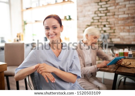 Smiling caregiver. Beautiful hard-working caregiver wearing uniform smiling while visiting pensioner