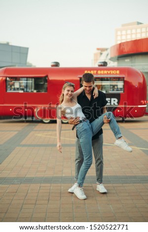 Funny couple young man and girl standing on street against red Food Truck background and having fun. Vertical photo of a fun couple. the guy wants to raise the girl.Love story of cheerful young people
