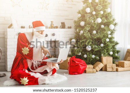 Modern Santa Claus. Cheerful Santa Claus working on laptop and smiling while sitting at his chair with fireplace and Christmas Tree in the background