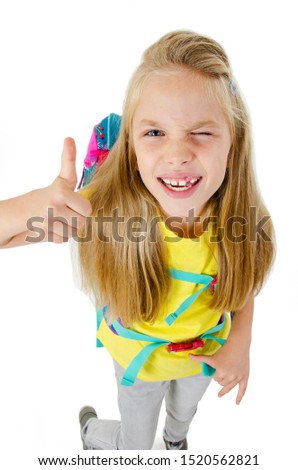 Cute little girl in casual school clothing is showing thumbs up sign. Winking and smiling. Isolated on white background. Wide angle.