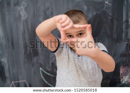 happy cute little boy having fun making hand frame gesture while standing in front of black chalkboard