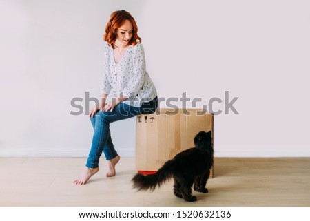 Young beautiful girl moving in the new apartment. Sitting on a carton box on a white background, black fluffy cat beside.