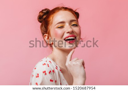 Beautiful woman with red hair covers her face with her hands. Girl in t-shirt with cherries cute smiles on pink background