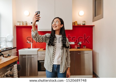 Joyful girl in striped shirt and glasses makes selfie on background of kitchen