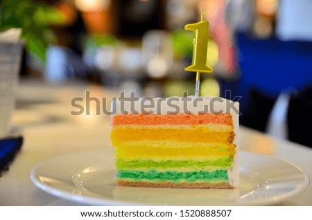 slice of rainbow cake with gold color number one candle on white plate. rainbow birthday cake 