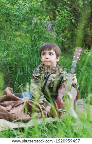 Young boy with guitar and backpack, traveler is resting in the forest and is playing with little black kitten.