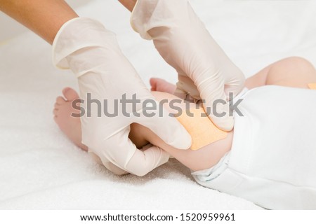 Doctor hands in white rubber protective gloves putting adhesive bandage on infant leg after injection of vaccine. Two month old baby. Medical concept. Closeup. Royalty-Free Stock Photo #1520959961