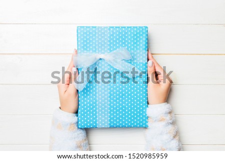 Female's hands holding striped gift box with colored ribbon on white rustic wooden background. Christmas concept or other holiday handmade present box, concept top view with copy space.