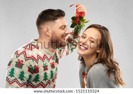 christmas, people and holiday traditions concept - portrait of happy couple in ugly sweaters kissing under the mistletoe over grey background