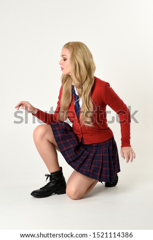 
full length portrait of blonde girl wearing red cardigan with tie and plaid skirt, school uniform. Sitting on the ground with  a white studio background.