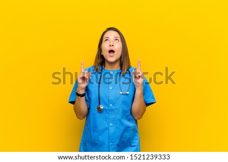 looking shocked, amazed and open mouthed, pointing upwards with both hands to copy space isolated against yellow wall