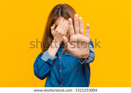 young pretty woman covering face with hand and putting other hand up front to stop camera, refusing photos or pictures against yellow background