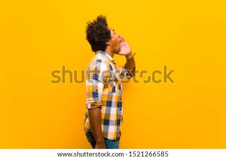 young black man profile view, looking happy and excited, shouting and calling to copy space on the side against orange wall