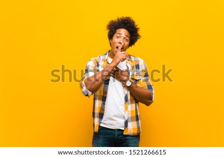 young black man looking serious and cross with finger pressed to lips demanding silence or quiet, keeping a secret against orange wall