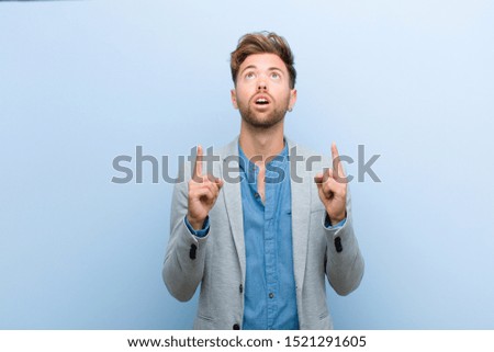 young businessman looking shocked, amazed and open mouthed, pointing upwards with both hands to copy space against blue background