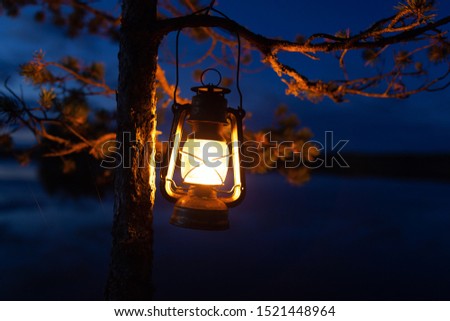 Vintage oil lamp hanging on a tree. Beautiful view of dark forest and lake at night. Hiker, Travel, Outdoor Concept