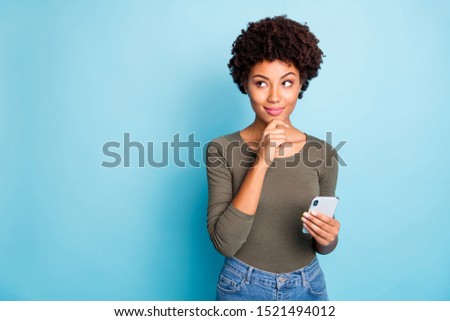 Photo of cunning smirking puzzled interested dark skinned youngster touching her chin in thought of what to post to her profile on social media wearing jeans denim isolated over vivid blue background