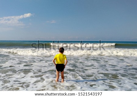 On a sunny day, a teenager went knee-deep into the water in the sea to meet the sea waves. A boy in a yellow T-shirt in black shorts. Photo taken from the back.