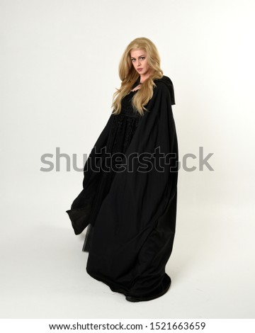 full length portrait of blonde girl wearing long black flowing cloak, standing pose  with  a white studio background.
