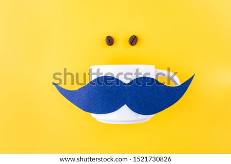 Blue paper mustache on a coffee mug, next to a coffee grain against a yellow background. Contrasting colors, minimalism, flat lay, top view. 