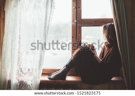 Pre teen child in warm woolen sweater seating on window sill and reading a book. Winter weekends in old log house. Cold snowy weather. Cozy homely concept. Royalty-Free Stock Photo #1521823175
