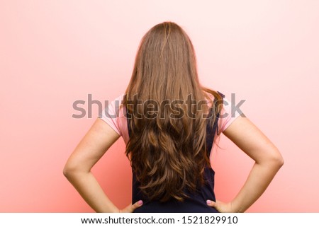 young  woman feeling confused or full or doubts and questions, wondering, with hands on hips, rear view against pink background