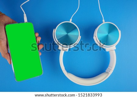 A hand holding a handphone with a green screen and blue white color headphone on a blue background