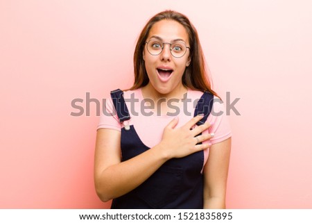 young  woman feeling shocked and surprised, smiling, taking hand to heart, happy to be the one or showing gratitude against pink background