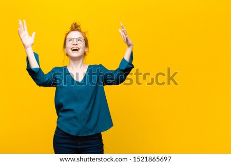 young red head woman feeling happy, amazed, lucky and surprised, celebrating victory with both hands up in the air against orange wall