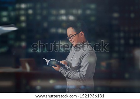 Young man looking happy and smiling while he’s reading a book. Student wearing long sleeves and eye glasses give positive emotion while reading a book. Reading and learning at midnight. 