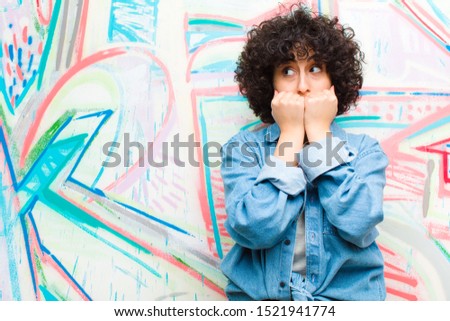 young pretty afro woman looking worried, anxious, stressed and afraid, biting fingernails and looking to lateral copy space against graffiti wall