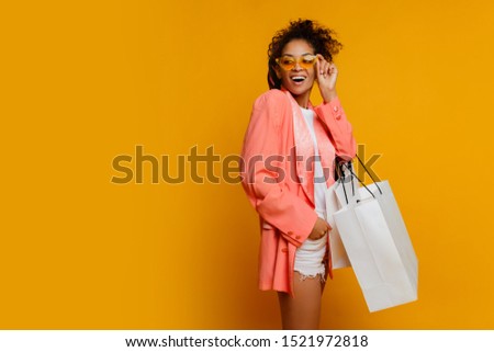 Fashionable elegant African female in pink costume holding white shopping bags on yellow background. Stylish curly hairstyle.