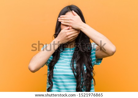 young pretty hispanic woman covering face with both hands saying no to the camera! refusing pictures or forbidding photos against brown wall