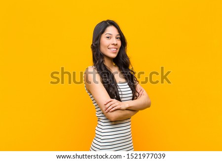 young pretty latin woman smiling to camera with crossed arms and a happy, confident, satisfied expression, lateral view against orange wall