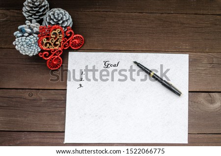 On a wooden background in the center lies a white sheet of paper with the words goals and numbers, with a fountain pen. In the upper right corner there are cones and a decorative carriage. Copy space