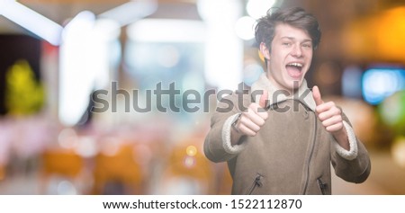 Young handsome man wearing winter coat over isolated background approving doing positive gesture with hand, thumbs up smiling and happy for success. Looking at the camera, winner gesture.