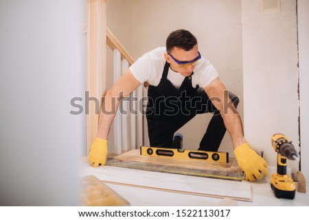 Maintenance man is  fixing the stairs. He is wearing glasses, yellow gloves and a black apron. A level measurement tool is next to him.
