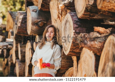 Beautiful woman in white sweater with maple leaves in hands stands on the background of firewood and logs and look to camera in autumn park. Closeup portrait.